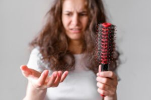 Concerned woman with medium-long hair holding a brush full of hair, indicating hair loss.