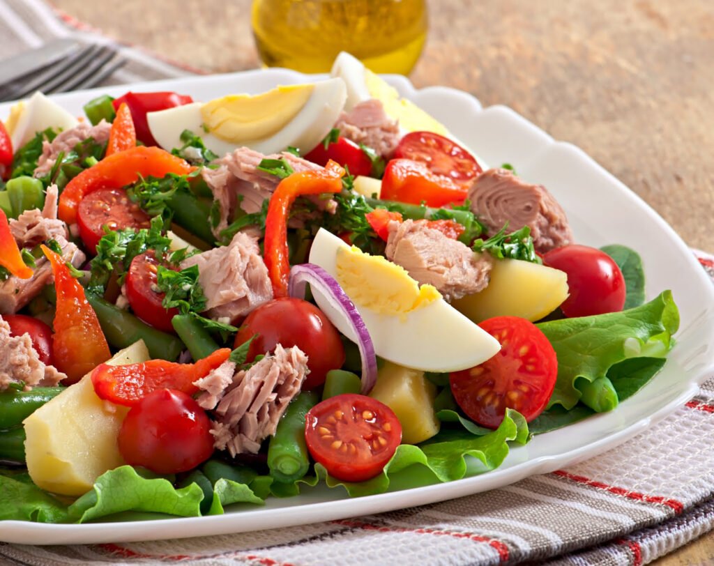 Plate of Mediterranean diet foods including salad, tuna, tomatoes, boiled eggs, and vegetables, with a glass jug of olive oil in the background.