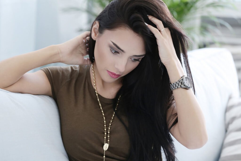 Indian woman with dark, healthy hair sitting on a sofa, placing her hand on her head to showcase her long, well-maintained hair.